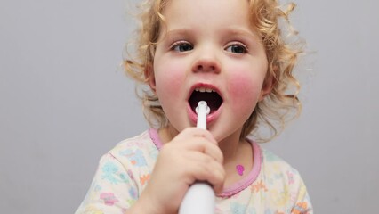 Child dental health. Baby's brushing routine. Electric toothbrush joy. Charming toddler baby with blonde wavy hair brushing teeth with electric toothbrush isolated over gray background