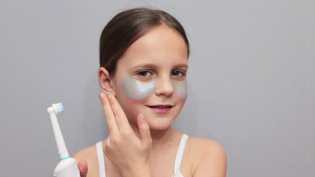 brown haired little girl applying under eyes patches taking care of her facial skin holding electric toothbrush for brushing teeth posing isolated on gray background