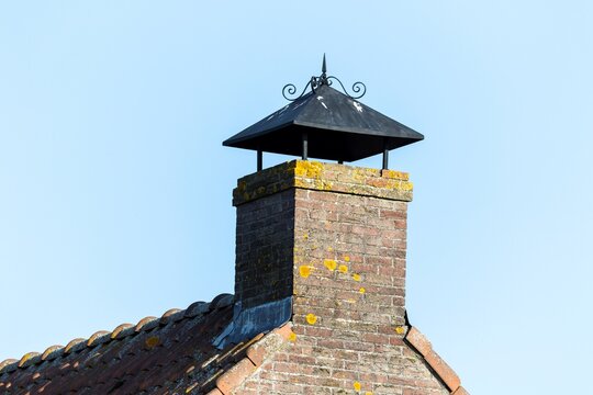 A portrait of a grey brick chimney with a small little roof build on top of it so the rain does not fall in it on a red tiled roof with a blue sky.