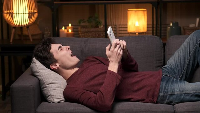 Young adult man lies on the sofa, engrossed in phone while letting out a yawn. The scene captures a relaxed evening, blending technology use and a hint of weariness in the comfort of living space.