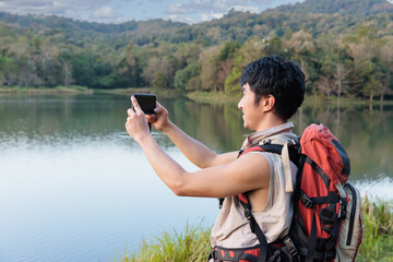 An Asian man trekking with backpack is smiling and happy making a picture of beautiful lake in a mountain using mobile phone during a sunny day