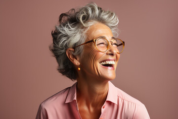 portrait of a beautiful elderly woman 60 years old in profile with a short haircut and glasses, laughs loudly, showing healthy teeth, in a pink blouse on a plain background in the studio