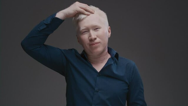 Portrait of an Asian albino guy. An eastern man with a genetic mutation of pigment touches his white hair on his head, corrects his hairstyle. The male is wearing a dark shirt in the studio