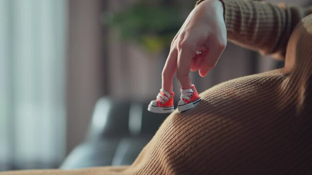 Pregnant woman walks her fingers in shoe over her large belly in living room at home, pregnancy, Pregnant female playing with her baby`s shoes.