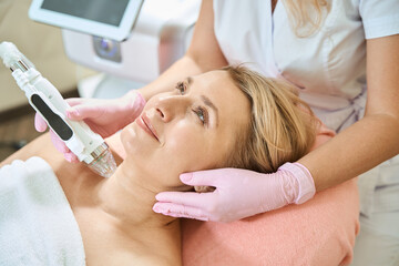 Cropped cosmetologist doing microneedle rf lifting of neck skin of smiling woman