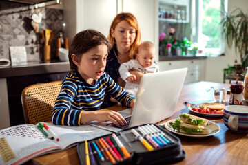 Mother multitasking with baby and son doing homework on laptop in kitchen