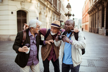 Three senior men laughing and sharing photos on a camera in a city square