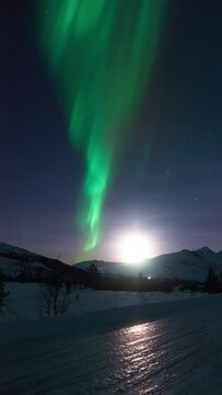 Aurora dancing elegantly next to a rising moon in a mountainous snowy landscape on January 1st 2024.