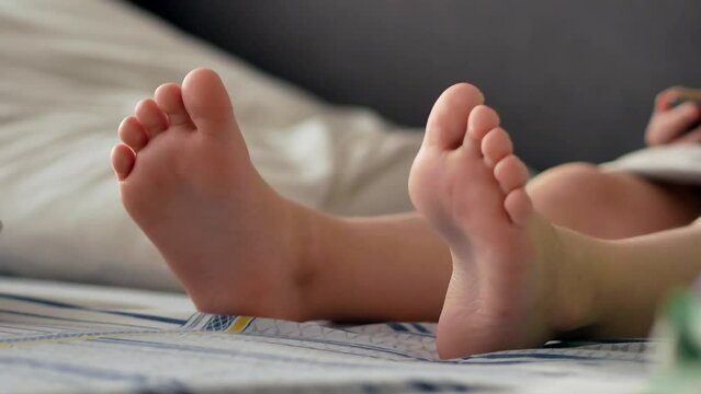The bare feet of a child sitting in bed in the morning with a smartphone, watching videos or playing games. Modern wireless technologies and the Internet.