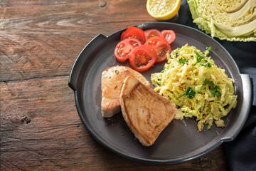 Fried tuna steaks with savoy cabbage and tomatoes on a dark plate and a rustic wooden table, copy space