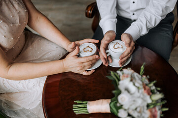 An adult man, a groom and a woman bride are sitting at a wooden table in a cafe holding cups of milk coffee and a foam heart in their hands. Wedding photography, food, ceremony.