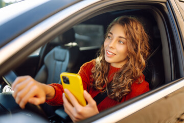Portrait of a young woman sitting in a car in the driver's seat looking into a smartphone, paying...