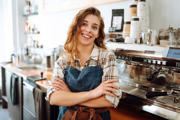 Portrait of a beautiful barista girl in an apron in a modern cafe bar. Startup successful small business owner  beauty woman stand in coffee shop restaurant. Business concept. - obrazy, fototapety, plakaty