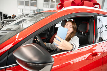 Woman making selfie while sitting in automobile in dealership buying new car