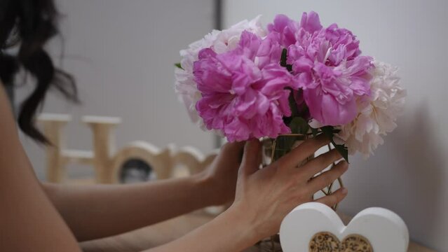 A girl puts pink and white peonies in a glass vase on a wooden nightstand in a modern living room in an apartment. A girl with long, black hair bends down and smells flowers. Close-up.
