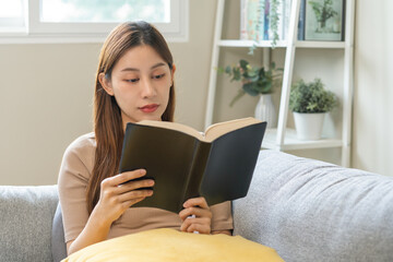 Having relaxing weekend, comfort asian young woman with cup of beverage, drinking coffee or tea of mug. Girl sitting on sofa open reading book enjoy of rest after get up in morning lifestyle at home.