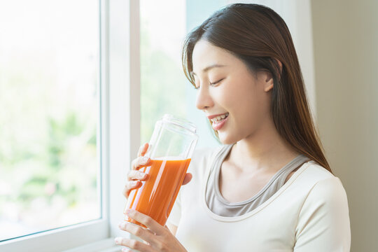 Orange detox juice, smile asian young woman hand holding glass of vegetable juice, carrot smoothie for diet near window at home, drinking healthy meal food for weight loss. Lifestyle, vegan nutrition.