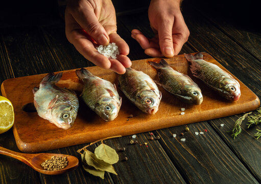 The cook adds salt to fresh fish before cooking. The process of salting crucian carp on the kitchen table with spices and coriander.