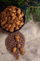 Top view of a deep black bowl filled with dried healthy almonds against the background of old burlap and a green branch of thuja. Delicious raw almond nut in a bowl against a burlap cloth background