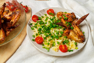 Farfalle pasta with grilled chicken wings on a white plate decorated with cherry tomatoes and green onions next to a dish with wings