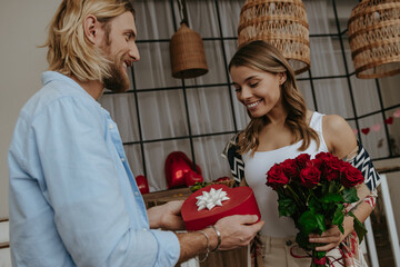 Young man giving a flower bouquet and gift box to his surprised girlfriend standing at home