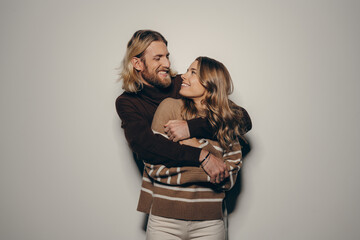 Joyful young loving couple in cozy warm wear embracing while standing on beige background