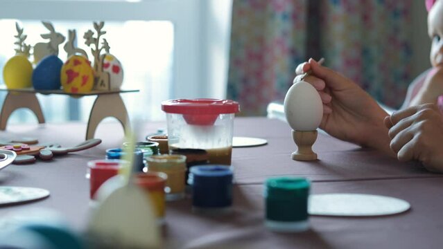 Close-up of hand of woman painting Easter egg at table by her baby eating cookies in the kitchen, selective focus