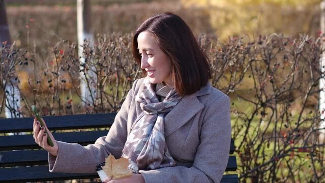 a cute fasion woman is sitting with mobile phone in autumn park. She is smiling.