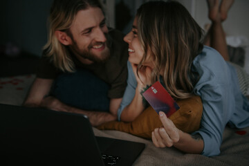 Joyful loving couple lying in bed while using laptop and credit card for online shopping from home