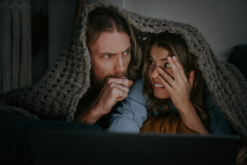 Scared young couple covering with blanket and watching horror movie on laptop while lying in bed
