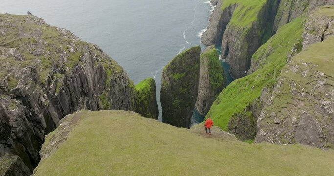 Aerial Shot Of Male Tourist Walking On Top Of Green Mountain, Drone Flying Forward Towards Birds Flying Over Sea - Faroe Islands, Denmark