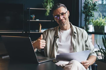 Poster Portrait of Caucasian male employee in earbuds holding paper documents and smiling at camera in office interior, successful businessman in classic spectacles posing at desktop with laptop computer © BullRun