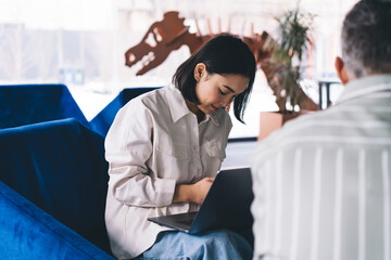 Asian woman with modern laptop computer communicate with unrecognizable male colleague during brainstorming meeting in coworking space, experienced colleagues discussing business and startup