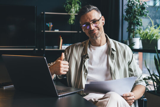 Portrait Of Caucasian Male Employee In Earbuds Holding Paper Documents And Smiling At Camera In Office Interior, Successful Businessman In Classic Spectacles Posing At Desktop With Laptop Computer