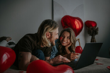 Joyful loving couple using laptop and shopping online at Valentines day from decorated bedroom