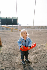 Little girl is standing near the swing, holding on to the seat