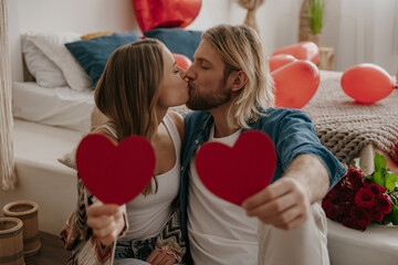 Loving couple embracing and kissing while holding heart shape Valentines cards at decorated bedroom