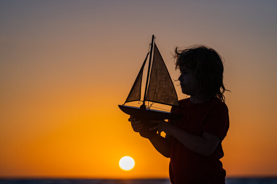 Silhouette of kid playing with toy seailing boat on sunset sea. Little boy playing with toy sailing boat, toy ship. Travel and adventure concept. Child feeling adventurous while cruising.