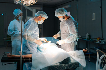 Wide shot of diverse team of professional male surgeons and female nurse performs invasive surgery on patient in hospital. African-American assistant helps head surgeon apply sutures.