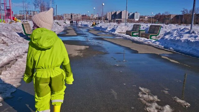 Rear view of cute child in green snowsuit walking down wet sidewalk with melting snow past muddy melt drifts.