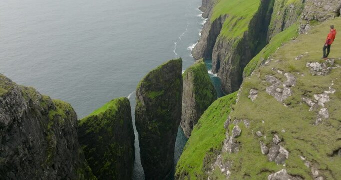 Aerial Panning Shot Of Male Tourist Standing On Top Of Faroe Island In Wavy Sea - Faroe Islands, Denmark