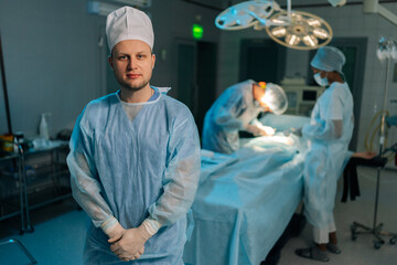 Medium shot portrait of male doctor in surgical uniforms standing posing looking at camera in dark modern operating room. Diverse team of surgeons performing operation on blurred background