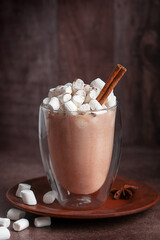 Traditional sweet hot chocolate or seasonal cocoa drink decorated with cinnamon stick and marshmallow topping served on plate with anise spice on dark brown table for winter festive breakfast