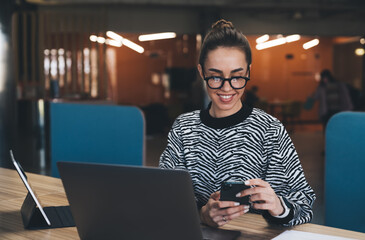 Cheerful young woman browsing smartphone while sitting with laptop and tablet in office
