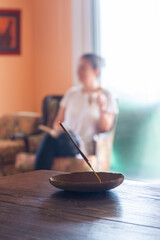 Incense stick burning in a cozy living room with a sunlit window in front of a blurred woman's...