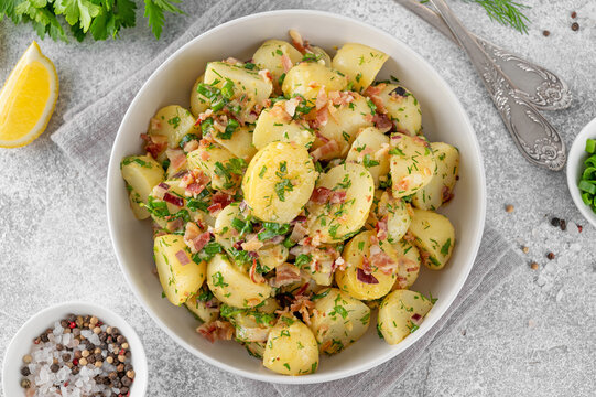 Warm potato salad with bacon, onion, fresh herbs and vinegar dressing in a bowl on a gray concrete background. German Kartoffelsalat.