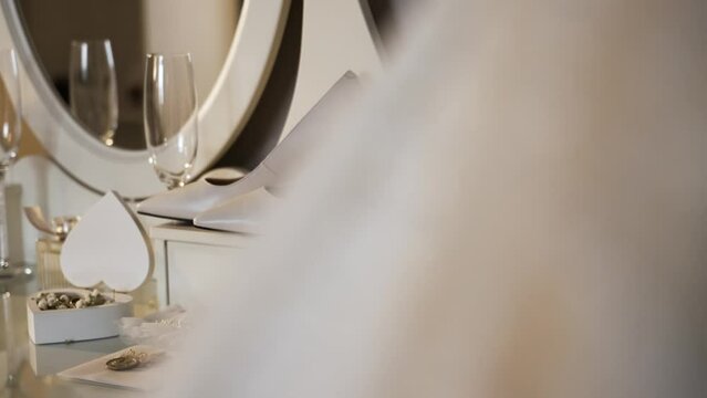 vanity setup with bridal accessories. There is a pair of elegant high-heeled shoes, a bottle of champagne, and two wine glasses on the table. A wedding dress with bead details hangs to the side