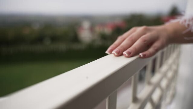 woman's hand gently resting on a white balcony railing, her nails well-manicured with a French tip design. The background is softly blurred, showcasing a distant landscape that suggests relaxation