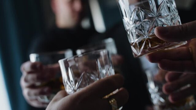 hand holding a glass of whiskey. The person is wearing a white shirt with a cufflink visible. Another hand with a glass is in the background. The focus is on the glass in the foreground, suggesting 