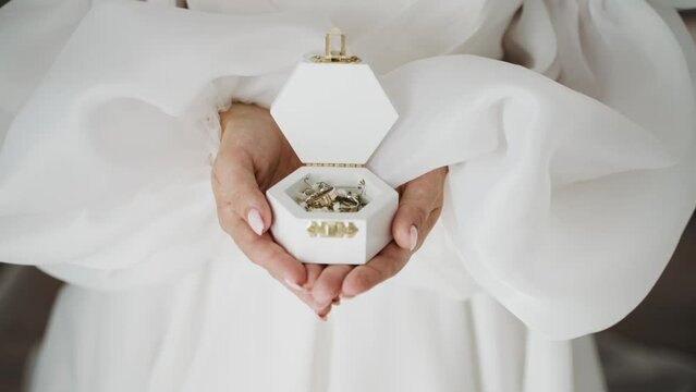 A bride holds a white hexagonal jewelry box with wedding bands, delicately captured against her flowing white gown, symbolizing wedding preparations.